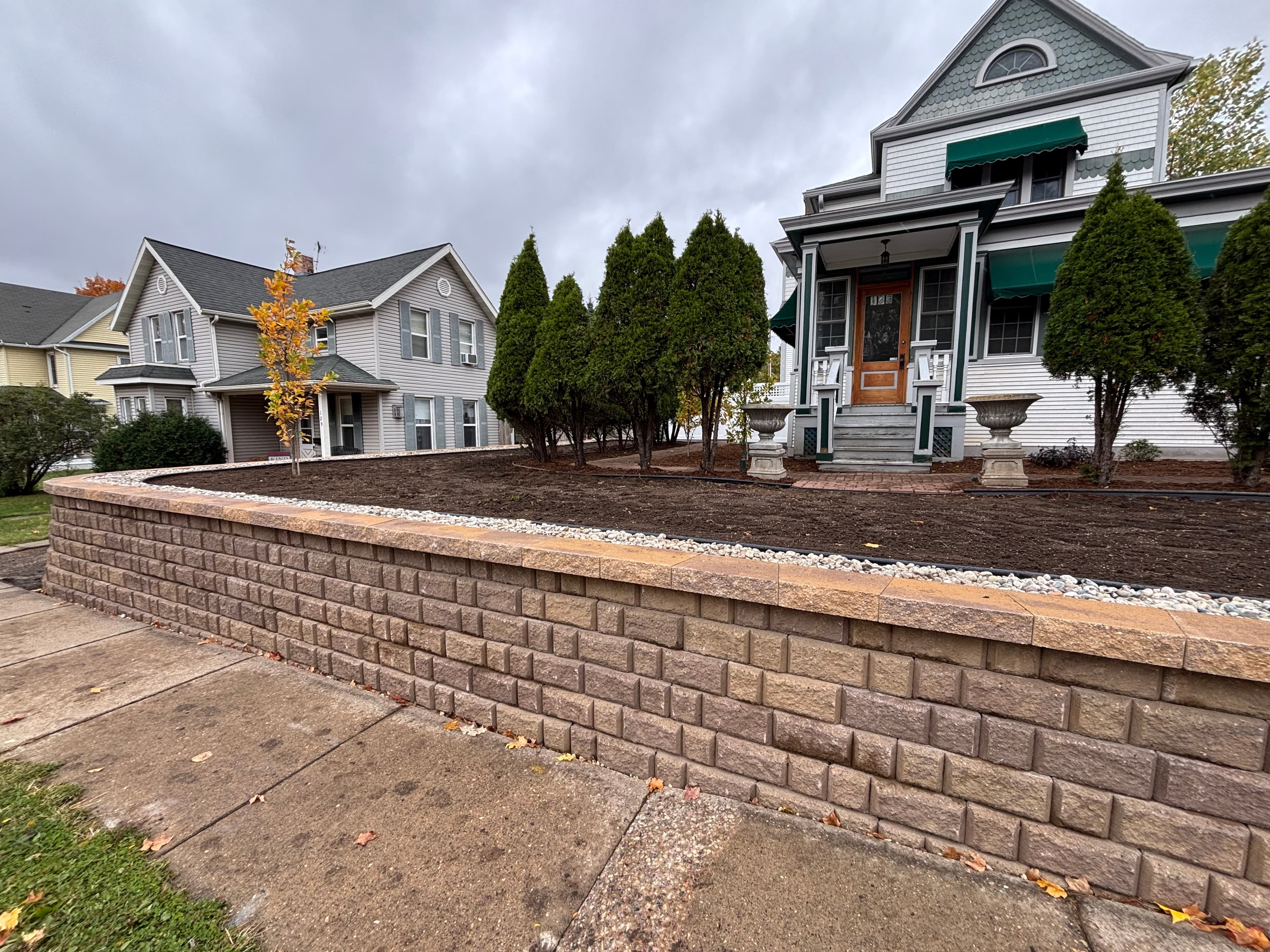 Terraced retaining wall landscaping with mature evergreen trees, fresh mulch installation, and decorative stone borders at historic Victorian home in Baraboo Wisconsin