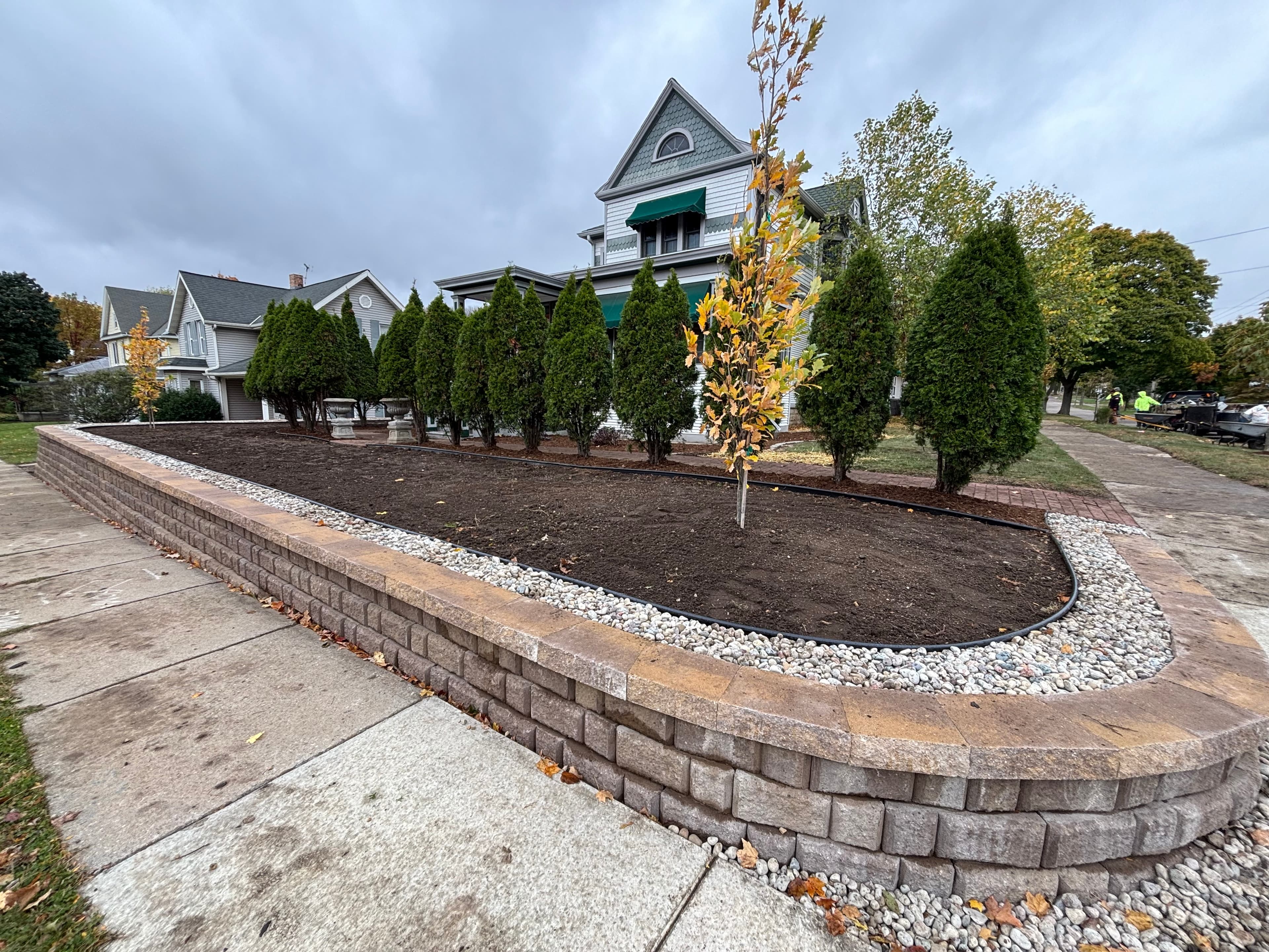 Curved segmental retaining wall with fresh green sod lawn, mature evergreen arborvitae privacy screening, and ornamental tree plantings in Baraboo WI residential landscaping