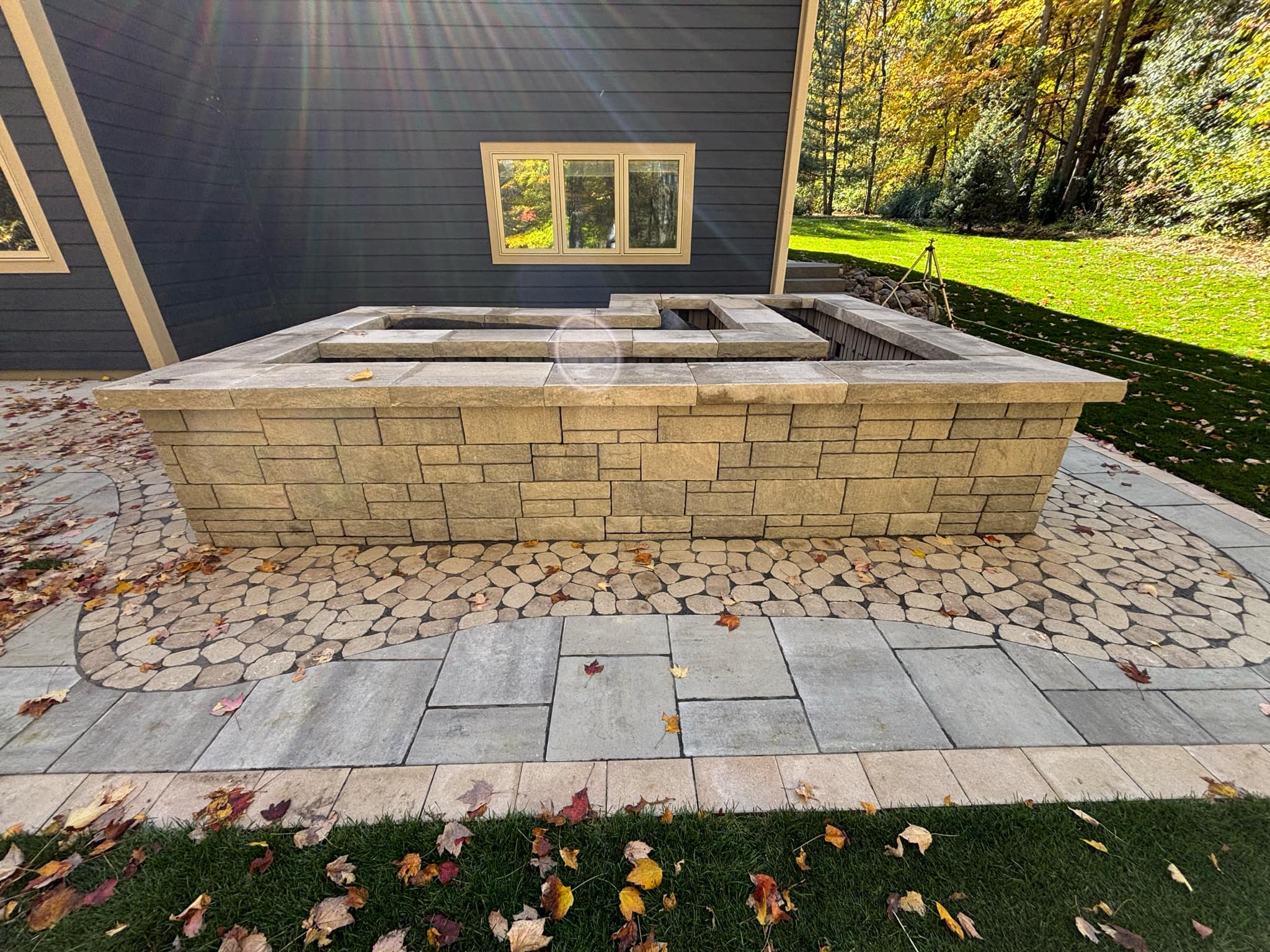 Close-up view of raised stone planter box with segmental retaining wall blocks in decorative pattern, flat stone cap countertop, two square openings on top, multi-textured paver patio with circular cobblestone pattern and large format pavers, dark gray house siding, professional craftsmanship Wisconsin