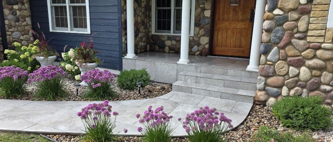 Front entrance with light gray concrete paver patio and steps, purple globe allium flowers, white and lime green hydrangeas, ornamental grasses, natural river rock stone veneer in mixed earth tones, navy blue siding, Oxford Wisconsin residential landscaping