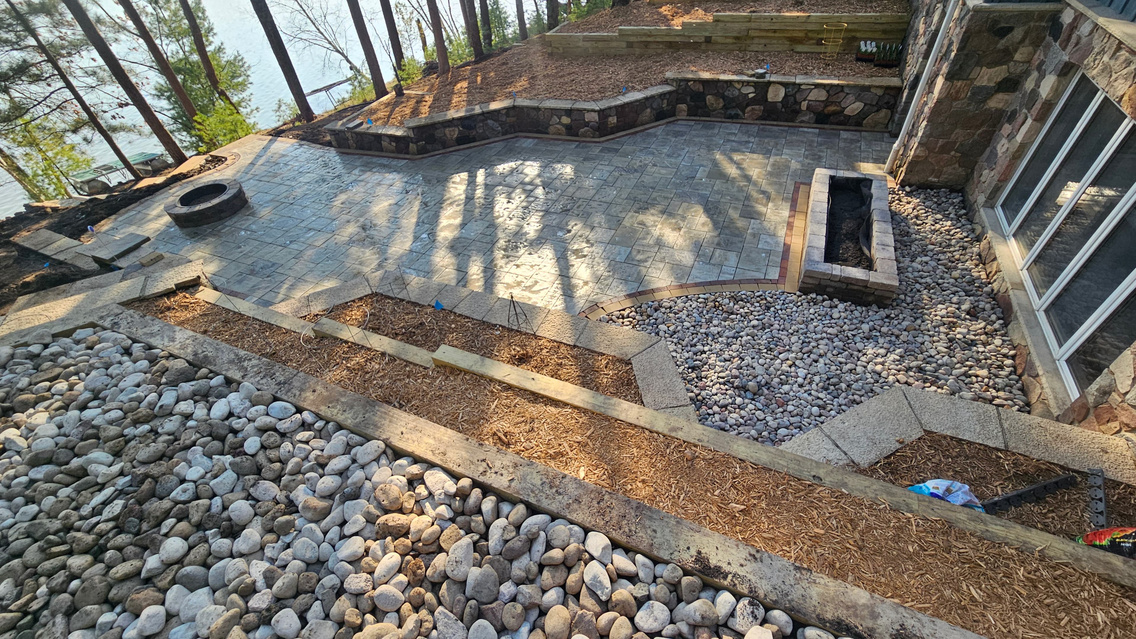 Aerial view of paver patio with accent borders, fire pit, river rock drainage beds, and timber retaining walls at lakefront property in Wautoma, WI