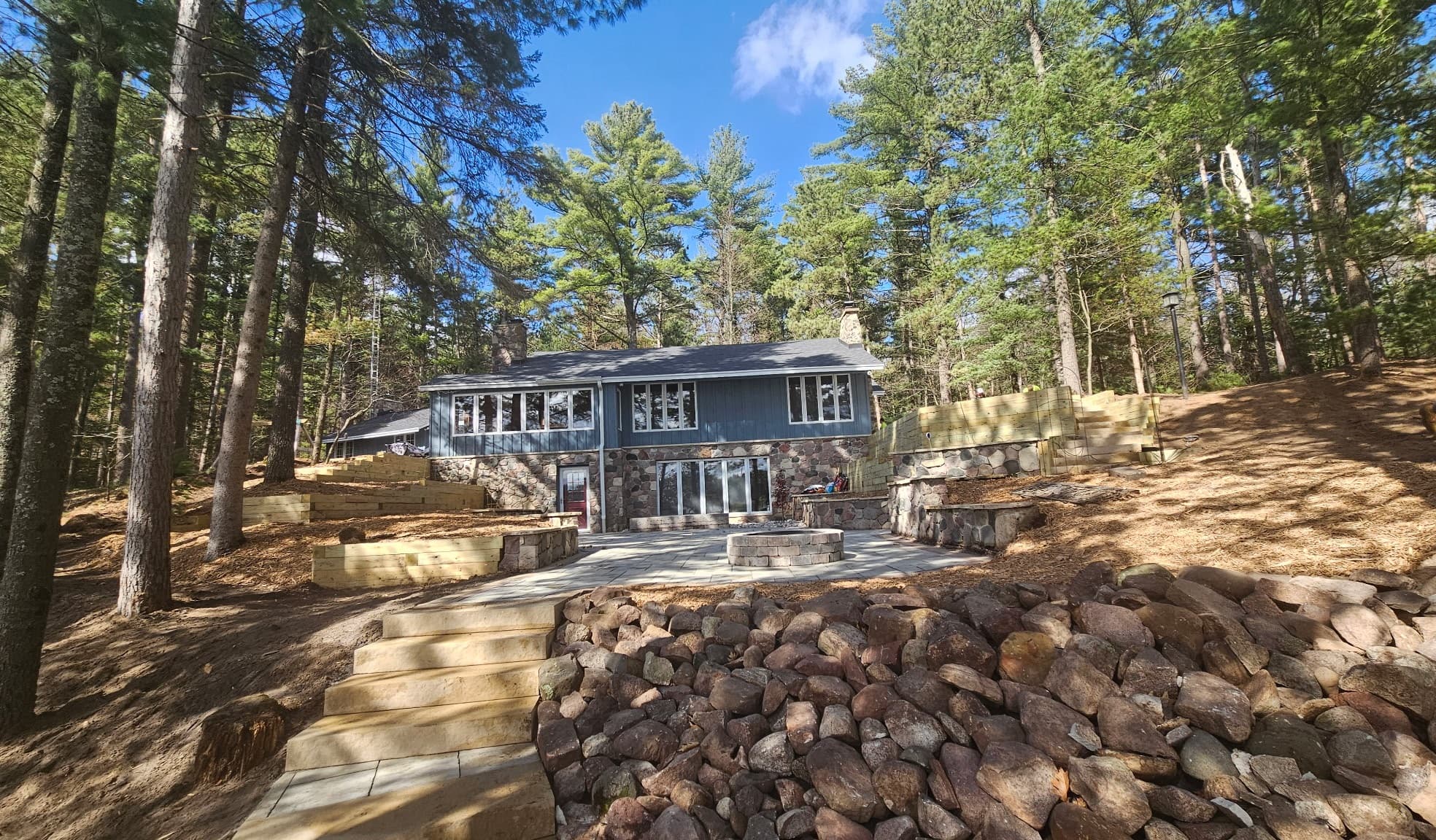 Wide view of paver patio with fire pit, natural limestone steps, boulder retaining walls, and wooded lakefront setting in Wautoma, WI