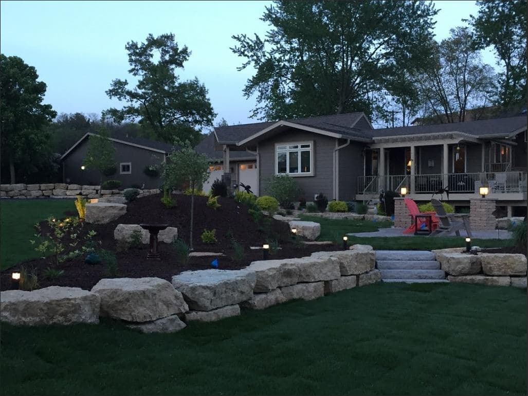 Professional residential landscaping at dusk featuring multi-tiered natural stone boulder retaining walls, landscape lighting, fresh sod installation, stone steps, ornamental plantings in dark mulch, Wisconsin Dells custom landscape design with terraced elevation changes